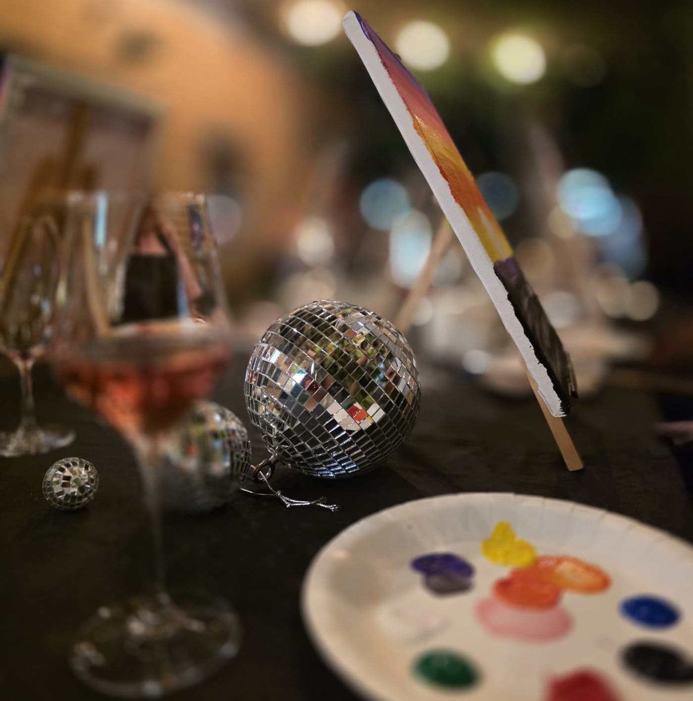 A disco ball, a paint palette and a glass of rosé beside a canvas.