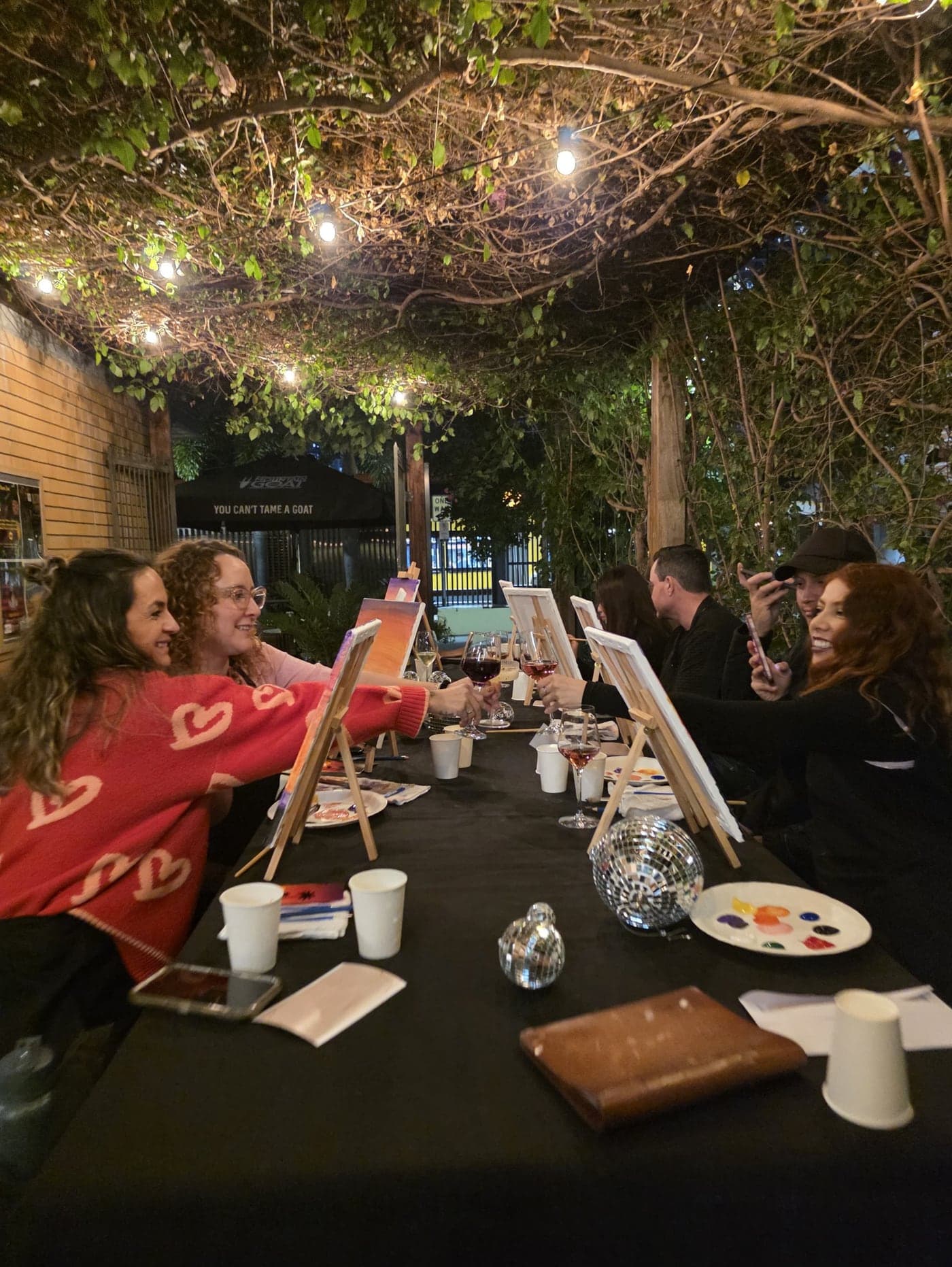 Friends painting at a long table under festoon lights and greenery.