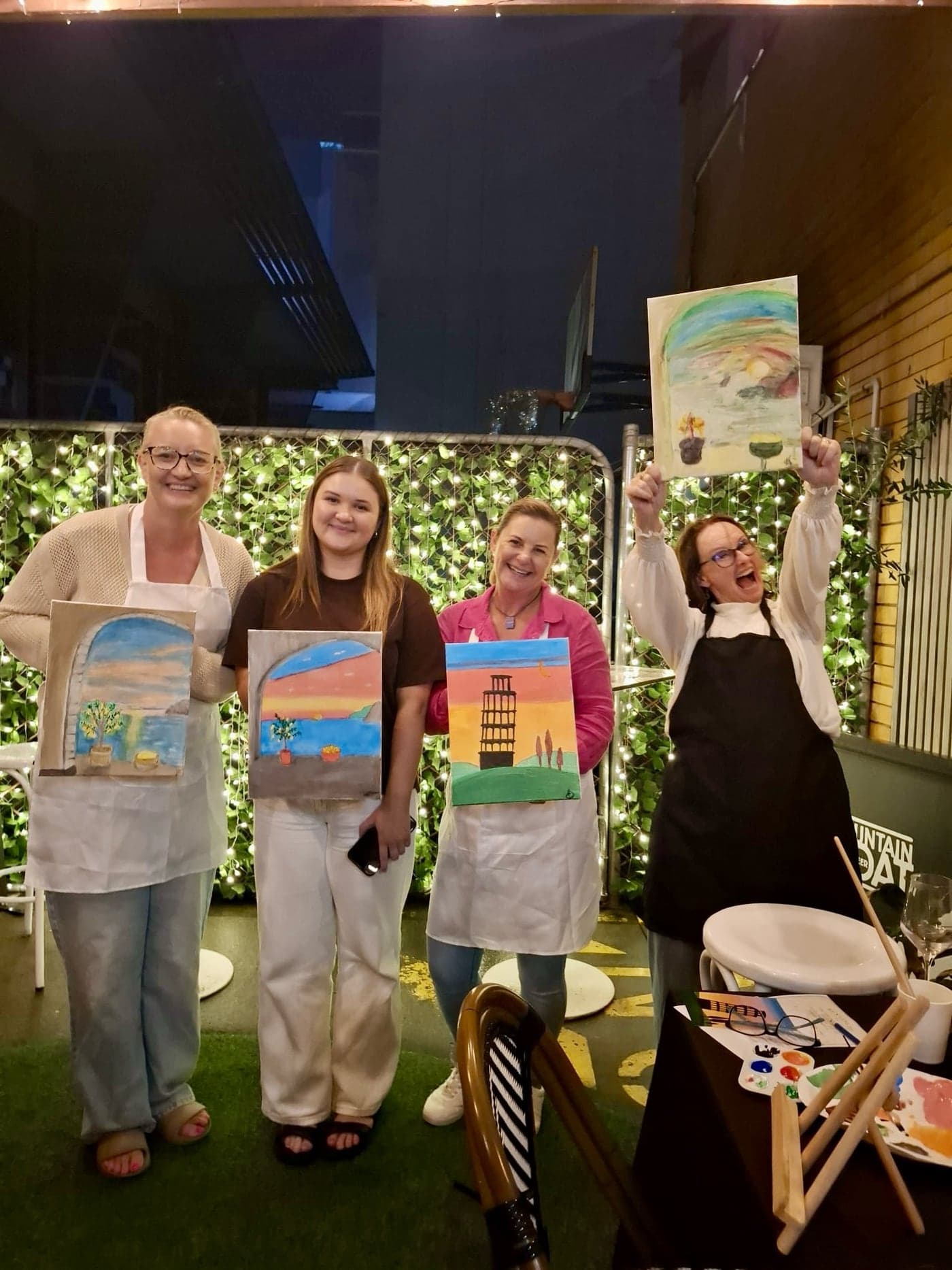 Four guests holding up their finished paintings in front of a green wall.