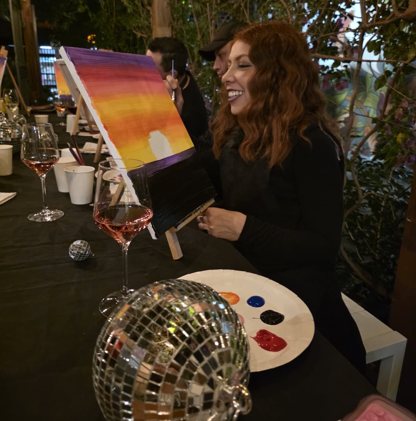 A guest smiling as she paints a sunset, a disco ball and a glass of rosé on the table.