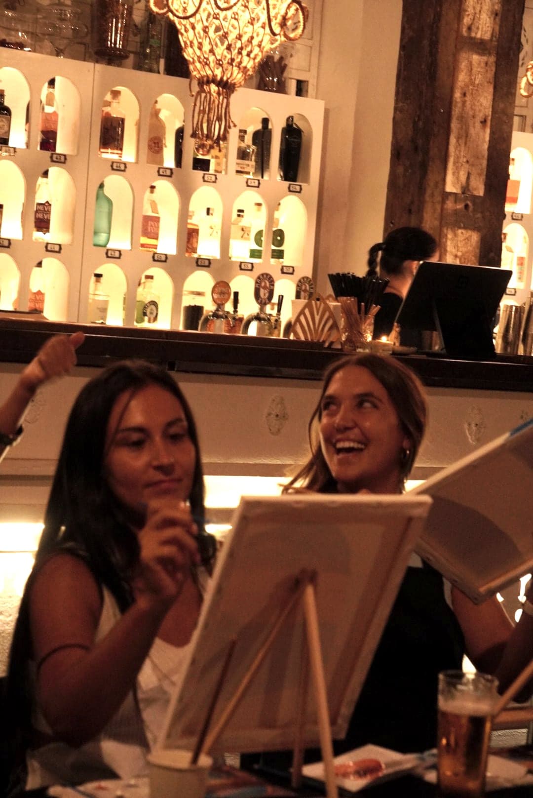 Two guests laughing over their canvases in front of a lit bar.