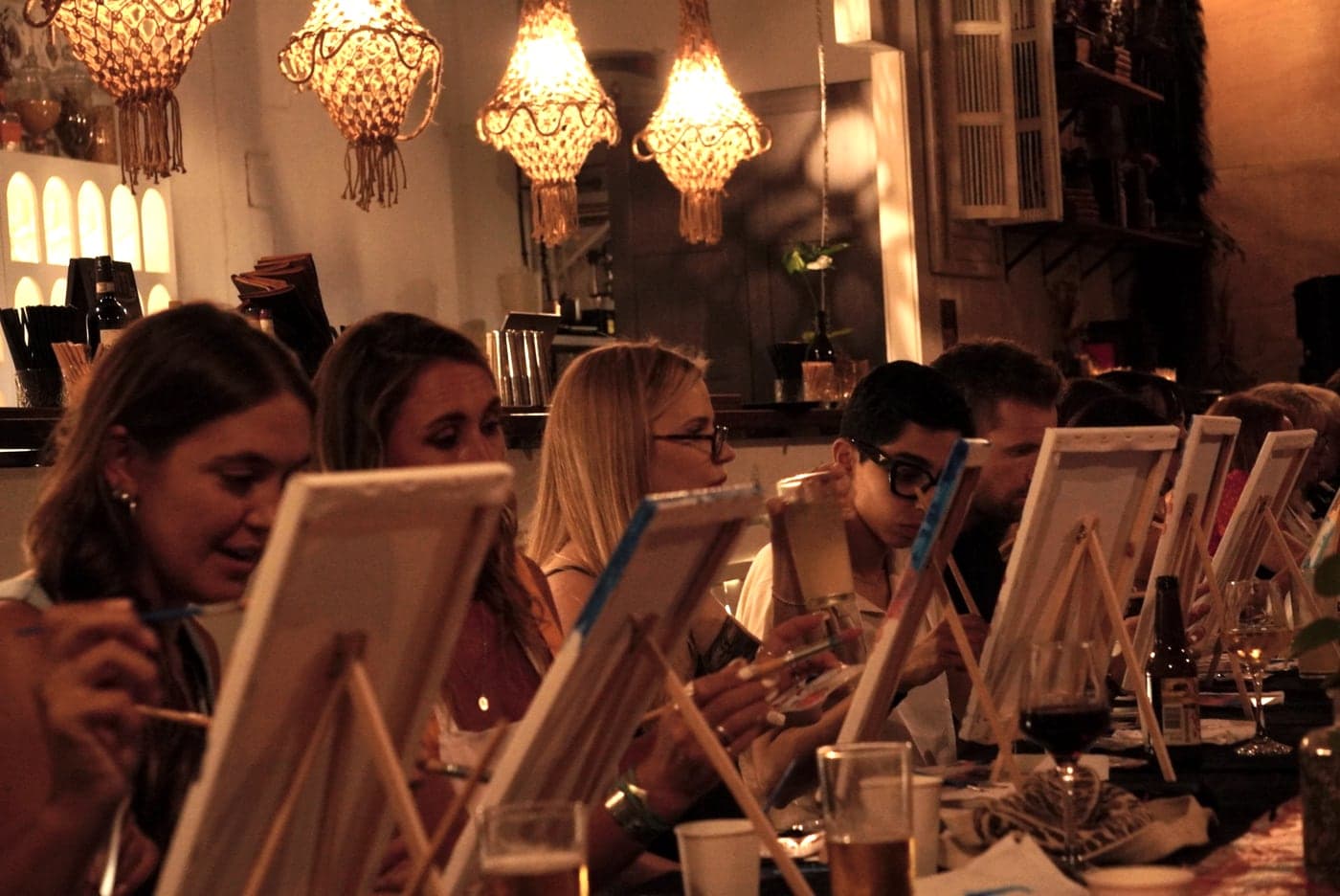 Guests painting at a long table under warm pendant lanterns.