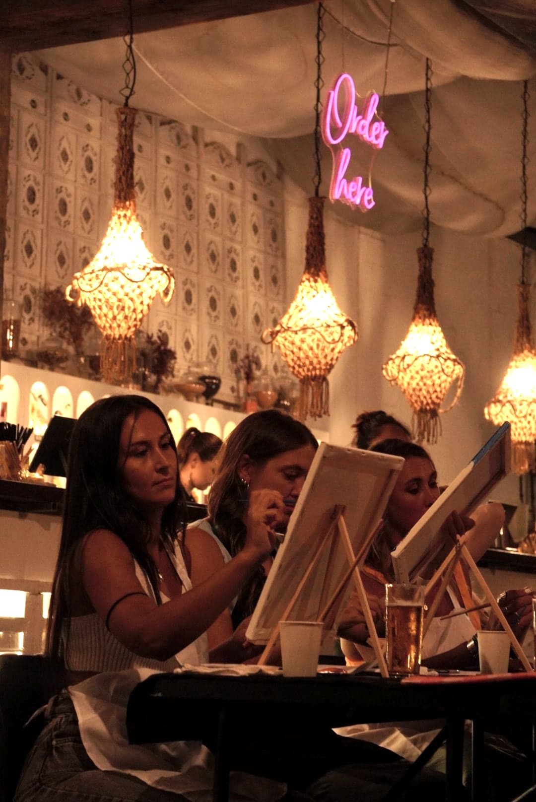 Guests painting under chandeliers and a pink neon sign.