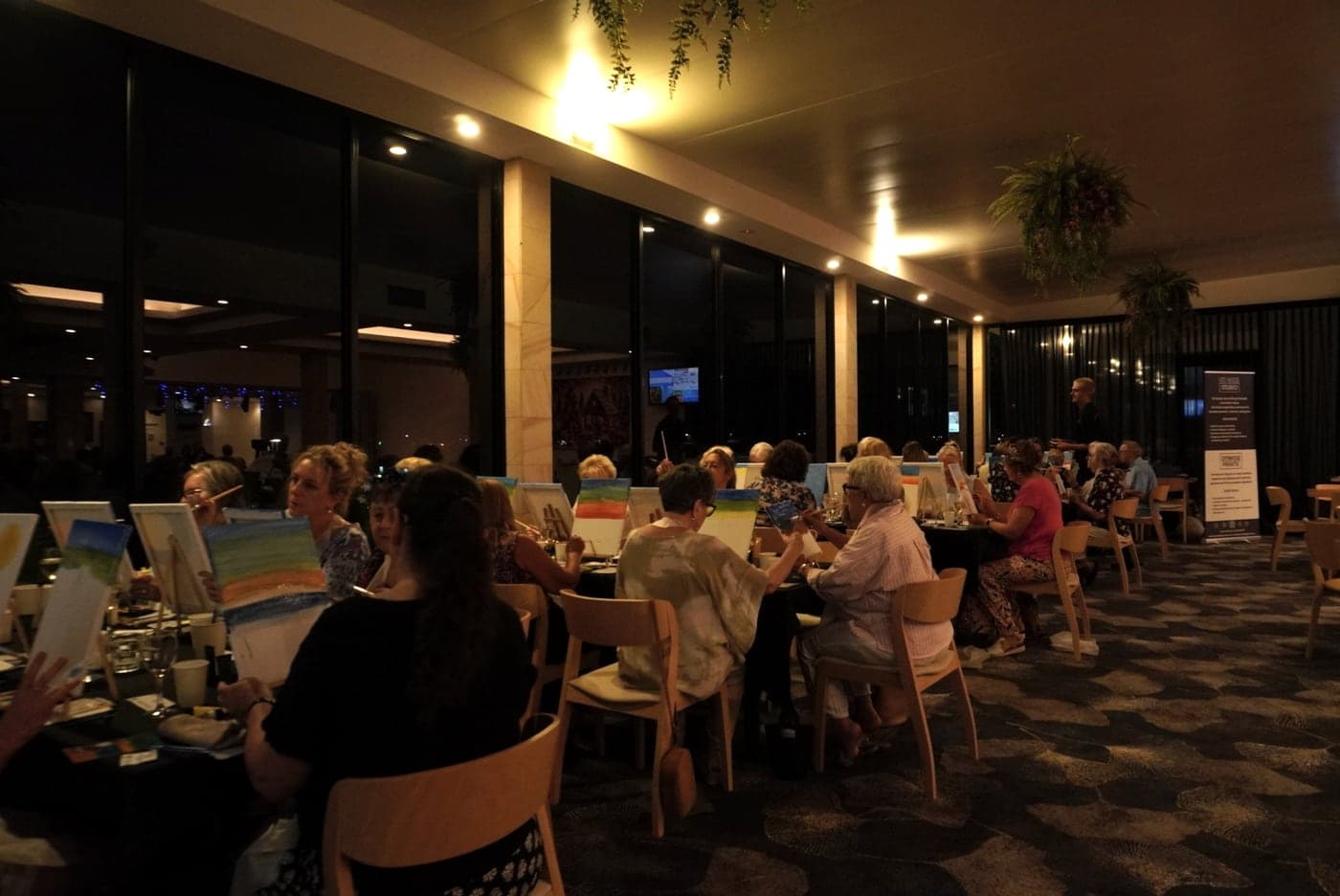 A full function room of guests painting at tables in the evening.