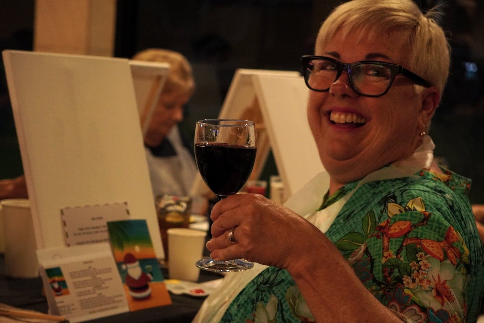 A smiling guest raises a glass of red wine beside her canvas.