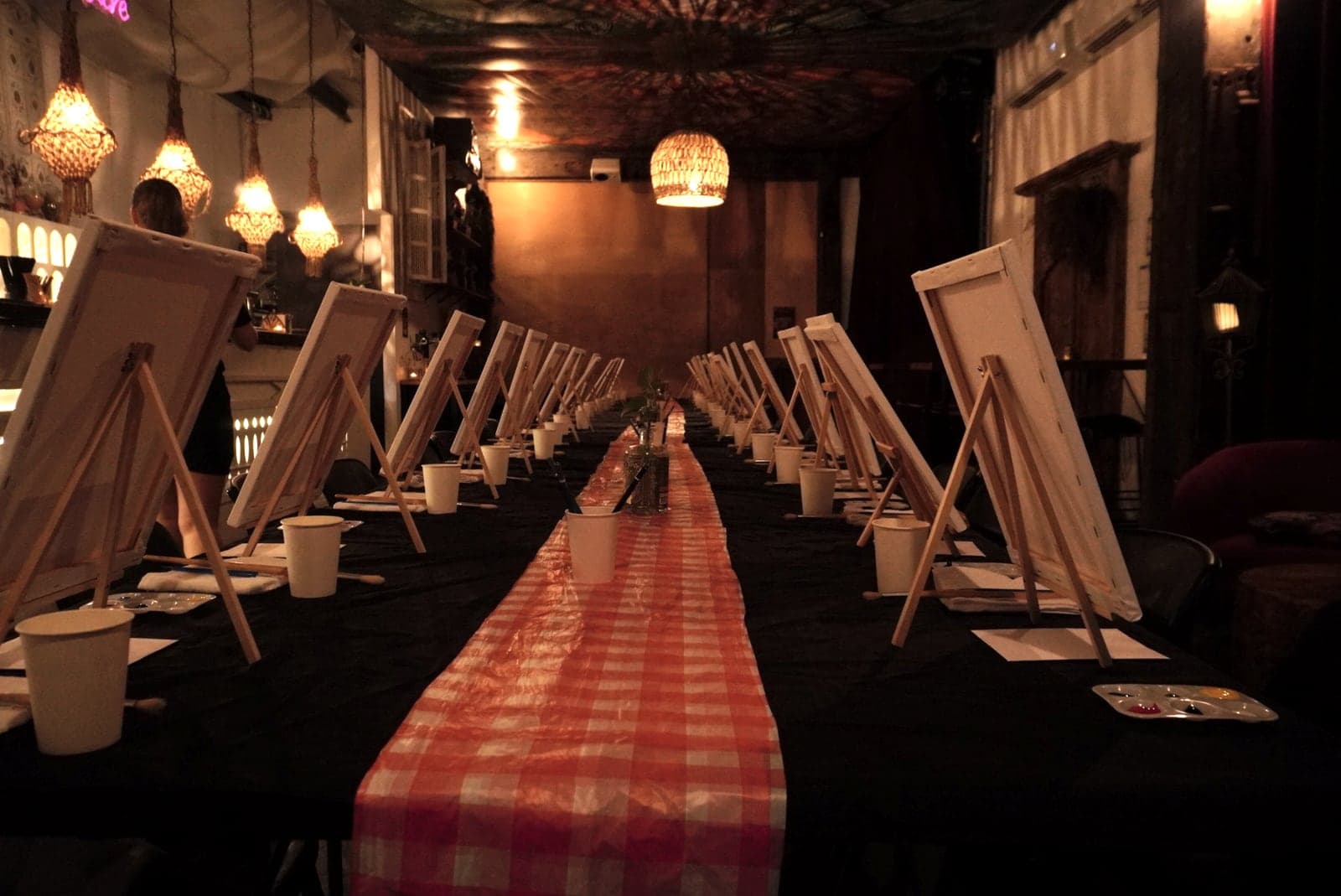 A long table set with easels, canvases and a red gingham runner before guests arrive.